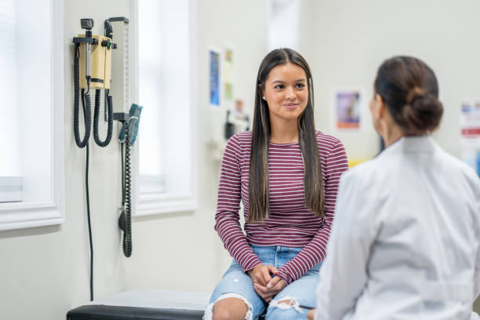 Young woman patient sits on an examination table in a clinic, speaking with a doctor; wall-mounted medical equipment is visible in the exam room.